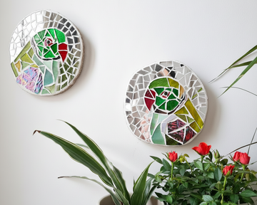 2 parakeet mosaics woth reflective silver mirror tiles displayed on a white wall with potted plants and flowers below