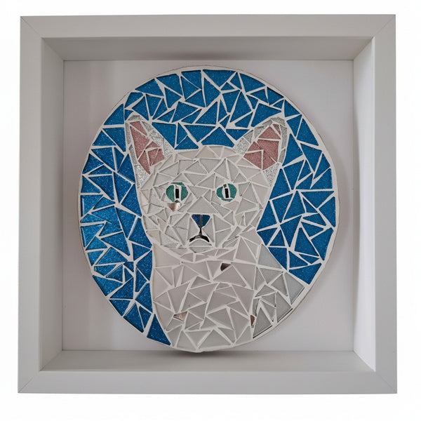 A circular mosaic wall art piece featuring a stylized white cat with blue and pink accents, mounted inside a square white shadow box frame.