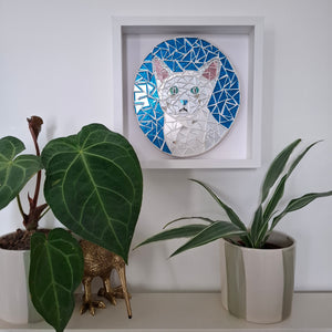 A framed mosaic art piece depicting a white cat with blue and pink details, mounted on a white wall above a shelf with potted green plants.