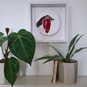A circular stained glass bird wall art piece featuring a robin, mounted in a square white shadow box frame on a white wall above a shelf with potted plants.