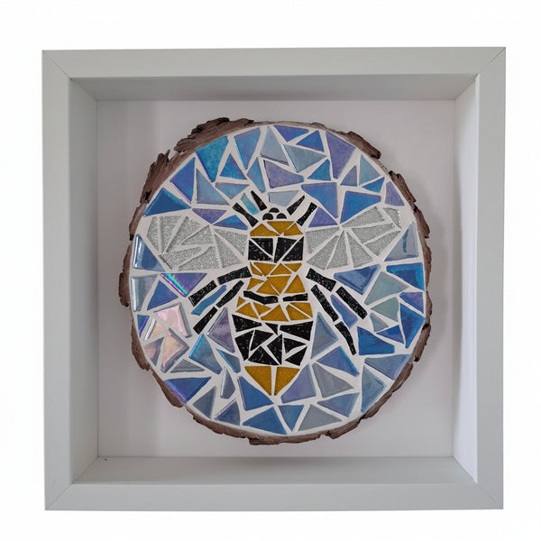 A square white shadow box frame containing a circular mosaic art piece featuring a stylized bee in yellow, black, and blue geometric shapes on a wood slice background.