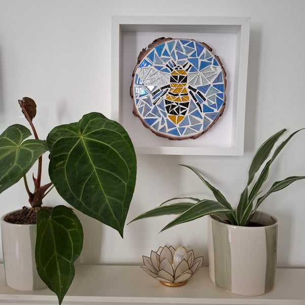 A square white framed piece of mosaic wall art featuring a stylized bee in blue, white, yellow, and black geometric shapes, mounted on a white wall above a shelf with potted plants.