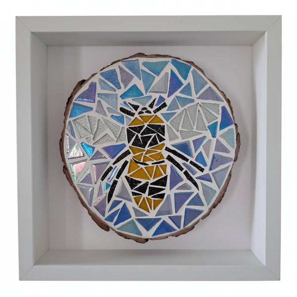 A square white shadow box frame displaying a circular mosaic art piece of a bee on a natural wood slice background.