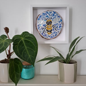 A square white-framed wall art piece featuring a colorful mosaic bee design on a natural wood slice, displayed on a shelf above potted green plants.