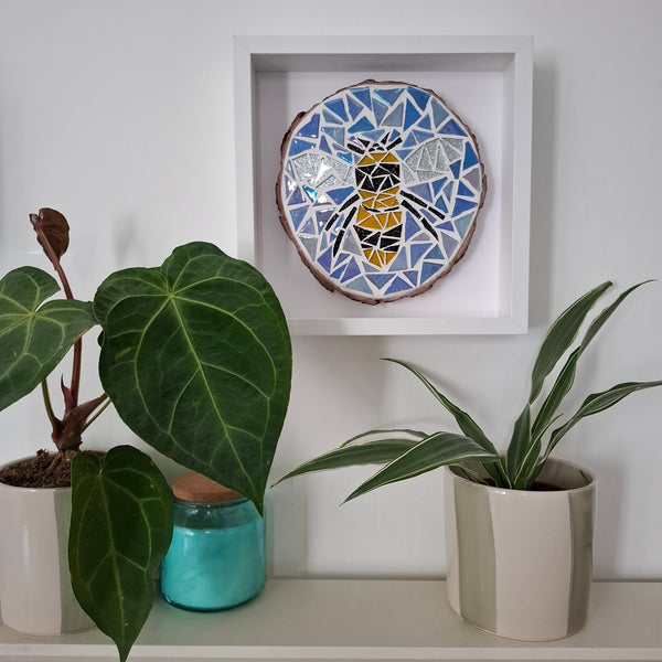A square white-framed wall art piece featuring a colorful mosaic bee design on a natural wood slice, displayed on a shelf above potted green plants.