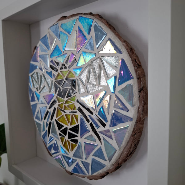 A round mosaic wall art piece mounted inside a white frame. The artwork features a yellow and black bee all set in white grout with a natural brown bark edge.