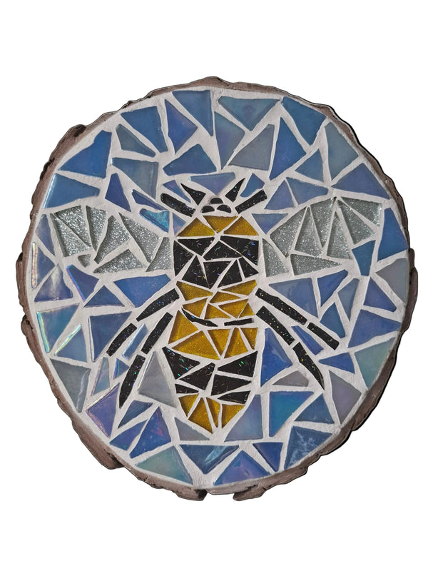 Circular mosaic wall art featuring a stylized bee design. Crafted with glittery black, gold and silver glass pieces on an iridescent blue background, set on rustic wood.