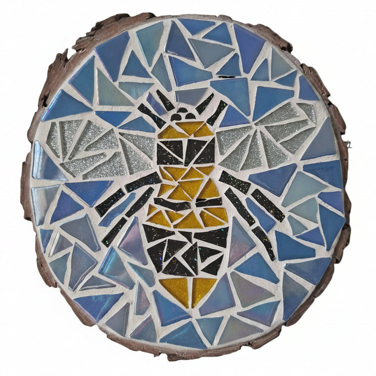 Circular mosaic wall art featuring a stylized bee design. Crafted with glittery black, gold and silver glass pieces on an iridescent blue background, set on rustic wood.
