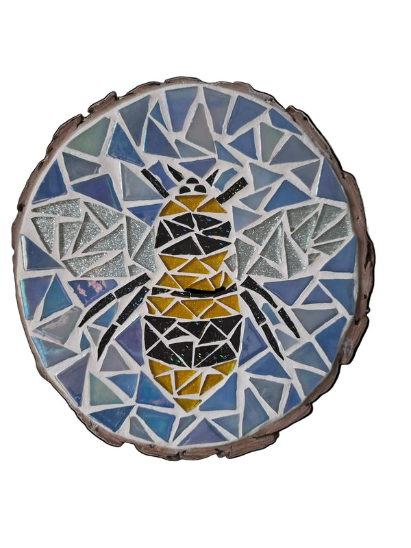 Circular mosaic wall art featuring a stylized bee design. Crafted with glittery black, gold and silver glass pieces on an iridescent blue background, set on rustic wood.