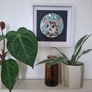 A framed mosaic art print of a dog's face hangs on a white wall above a shelf with plants and a glass bottle. The circular mosaic features geometric shapes in teal, brown, beige, and white, set against a dark background inside a white frame.