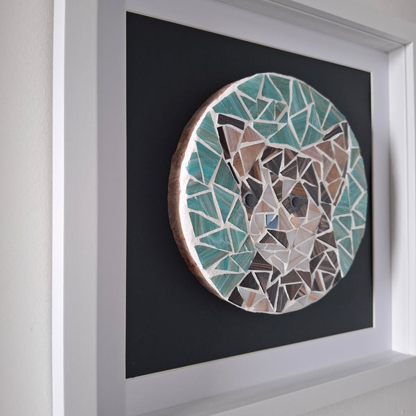 A framed mosaic wall art piece depicting a dogs face, made from teal, brown, beige, and white geometric tiles on a dark background, housed in a white frame.