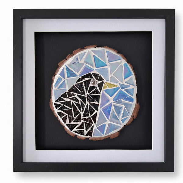 A square black-framed wall art piece featuring a circular mosaic bird design on a dark background. The mosaic includes blue, light blue, black, white, and a small gold accent.