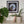 A framed circular mosaic bird wall art piece hangs on a white wall above a shelf with potted plants and a book. The artwork features a geometric bird design in blue, white, black, and brown tones inside a black frame.