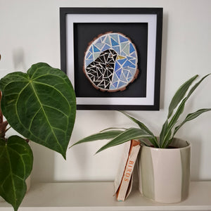 A framed circular mosaic bird wall art piece hangs on a white wall above a shelf with potted plants and a book. The artwork features a geometric bird design in blue, white, black, and brown tones inside a black frame.