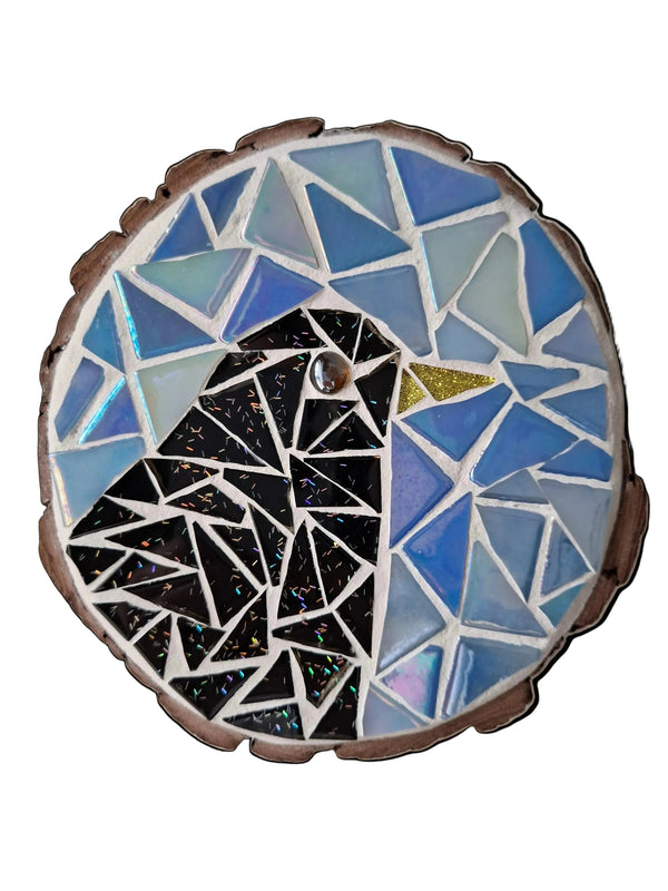Circular mosaic wall art featuring a stylized bird design. Crafted with glittery black glass pieces on an iridescent blue background, set on rustic wood.