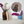 A styled interior shelf scene with a round blackbird mosaic wall art piece mounted on a white wall, alongside an amber glass vase with dried white and purple flowers, a blue ceramic pot with purple flowers, and a vintage book.