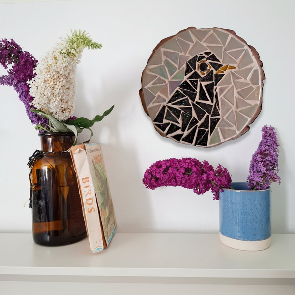 A styled interior shelf scene with a round blackbird mosaic wall art piece mounted on a white wall, alongside an amber glass vase with dried white and purple flowers, a blue ceramic pot with purple flowers, and a vintage book.