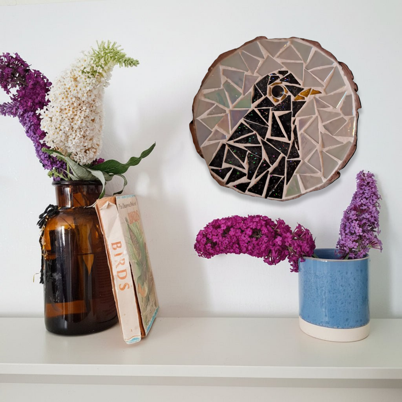 A styled interior shelf scene with a round blackbird mosaic wall art piece mounted on a white wall, alongside an amber glass vase with dried white and purple flowers, a blue ceramic pot with purple flowers, and a vintage book.
