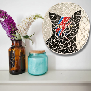 A circular mosaic wall art piece featuring a black cat with a David Bowie-style red, blue, and yellow face, displayed on a white wall above a shelf with an amber vase of flowers and a blue glass jar.