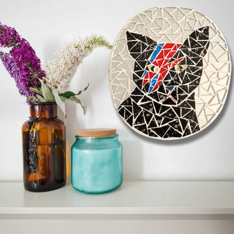 A circular mosaic wall art piece featuring a black cat with a David Bowie-style red, blue, and yellow face, displayed on a white wall above a shelf with an amber vase of flowers and a blue glass jar.