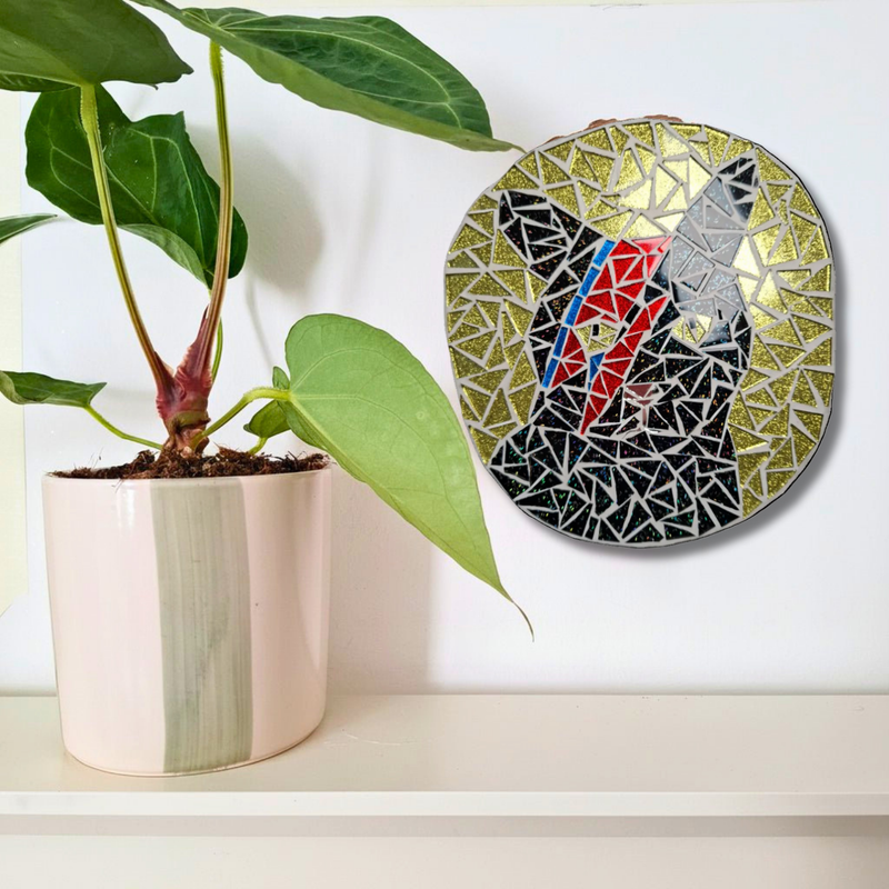 A round mosaic wall art piece featuring a stylized black cat with red, blue, and gold details, displayed on a white wall above a shelf next to a potted green plant.