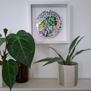 A circular mosaic wall art piece featuring a stylized green and red bird, displayed in a white square frame on a white wall above a shelf with potted plants.