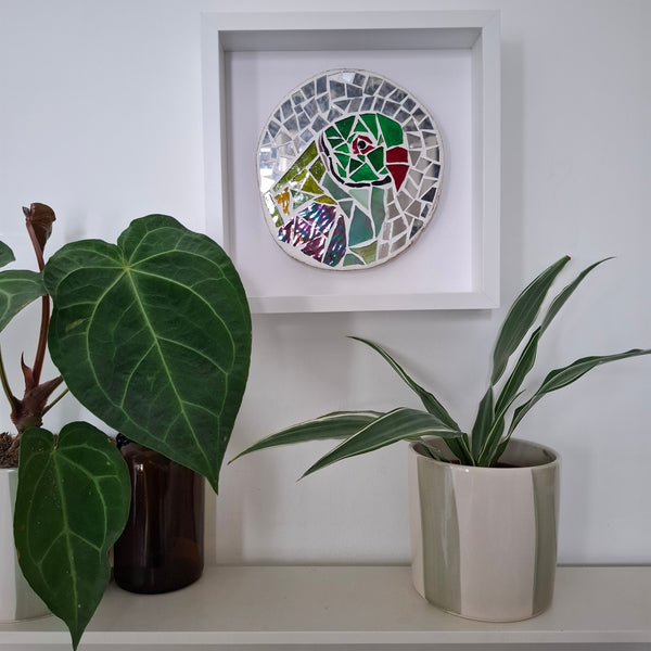 A circular mosaic wall art piece featuring a stylized green and red bird, displayed in a white square frame on a white wall above a shelf with potted plants.