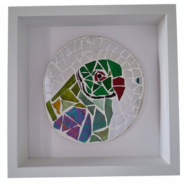 A circular mosaic wall art piece featuring a stylized parrot in green, red, purple, and yellow tones, mounted inside a white square shadow box frame.