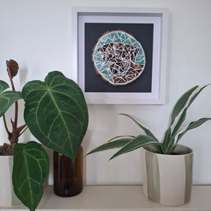 A square framed piece of mosaic art featuring a dog in teal, brown, and white on a dark background, displayed on a shelf above potted plants.