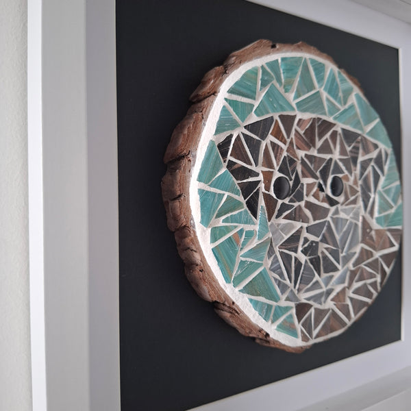 A circular mosaic wall art piece mounted on a dark square backing, framed in white. The artwork features a dog of teal, brown, grey, and white tiles