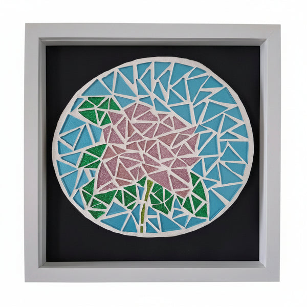 A square framed mosaic wall art piece featuring a stylized flower in pink and green with blue and light blue geometric petals, mounted on a black backing inside a white frame.