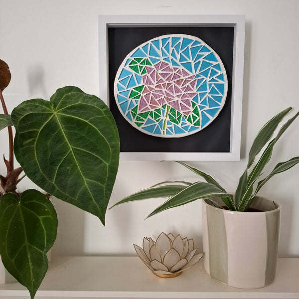 A framed circular mosaic wall art piece featuring a pink rose on a light blue background, displayed on a shelf above potted plants.