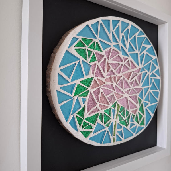 A circular mosaic wall art piece featuring a stylized flower in pink and green, set within a white frame with a black backing board.