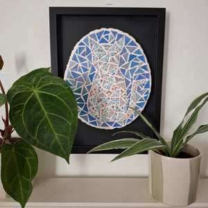A framed mosaic art piece depicting a cat in shades of blue, white, and red, displayed on a shelf next to green potted plants.