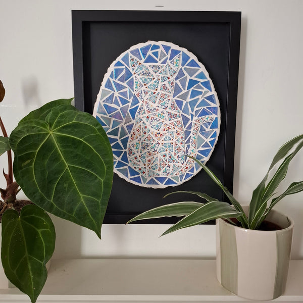 A framed mosaic art piece depicting a cat in shades of blue, white, and red, displayed on a shelf next to green potted plants.