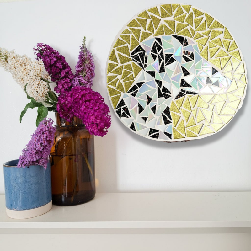 A round mosaic wall art piece with a dalmation, displayed on a white shelf next to two vases with purple and white flowers against a white wall.