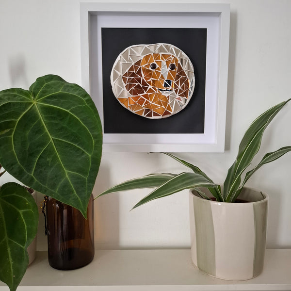 A framed geometric mosaic art piece depicting a dog's face in brown and bronze mirror tiles, styled on a shelf with potted plants against a white wall.