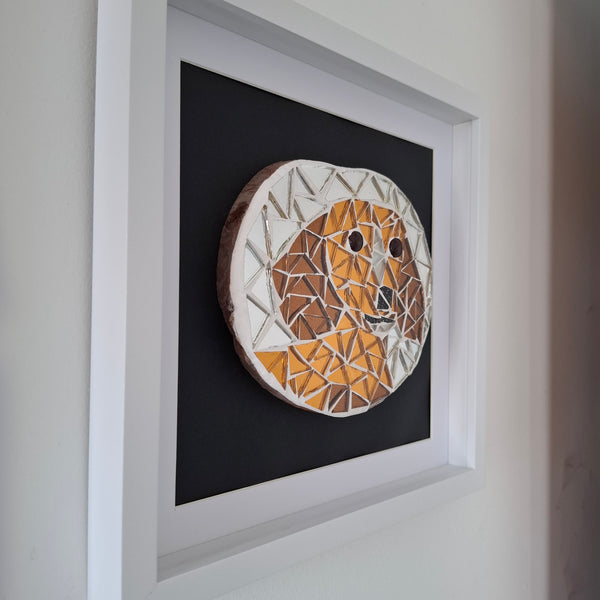 A framed circular mosaic wall art piece featuring a stylized dog face made of mirror tiles in bronze, brown, and white, mounted on a black background inside a white frame.