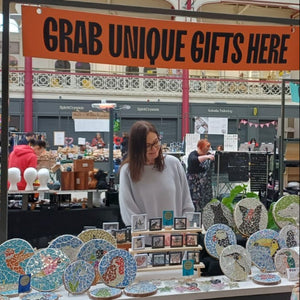 A market stall at an indoor fair with a large orange sign reading 'GRAB UNIQUE GIFTS HERE.' Artist Naomi Hope stands behind a display of colourful mosaic artwork of animals and birds