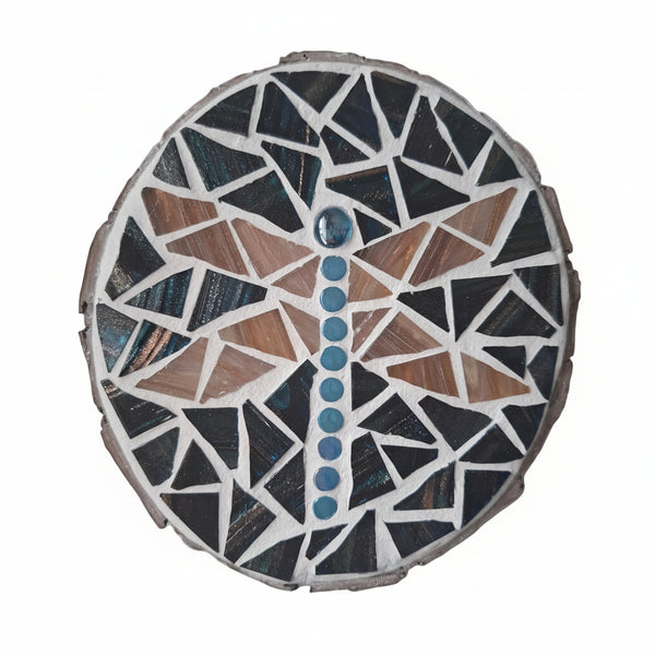Circular mosaic wall art featuring a stylized dragonfly design. Crafted with gold-dusted pink glass pieces and iridescent gems on a gold-dusted dark blue background, set on rustic wood.