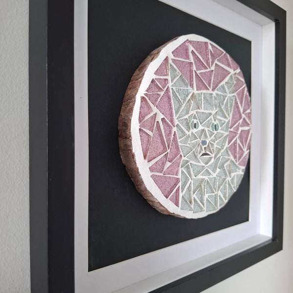 A framed circular mosaic wall art piece featuring a stylized cat face in pink, white, and pale blue geometric segments, mounted on a dark backing inside a black frame.