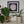 A framed circular mosaic art piece featuring a cat face in pink, white, and silver tiles, displayed on a shelf above green potted plants and a gold kiwi bird figurine.