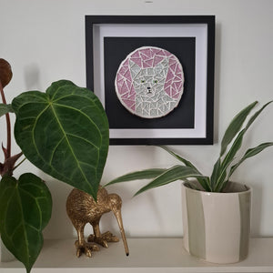 A framed circular mosaic art piece featuring a cat face in pink, white, and silver tiles, displayed on a shelf above green potted plants and a gold kiwi bird figurine.