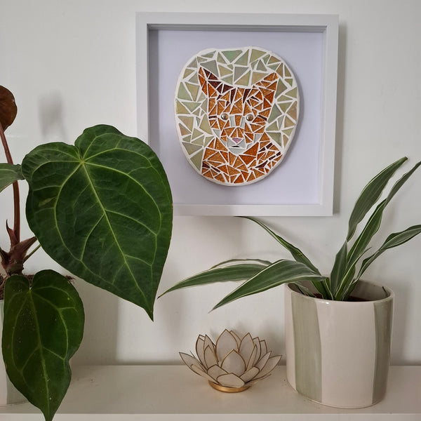 A framed mosaic art print of a cat's head hangs on a white wall above a shelf with potted plants and a decorative lotus candle holder.