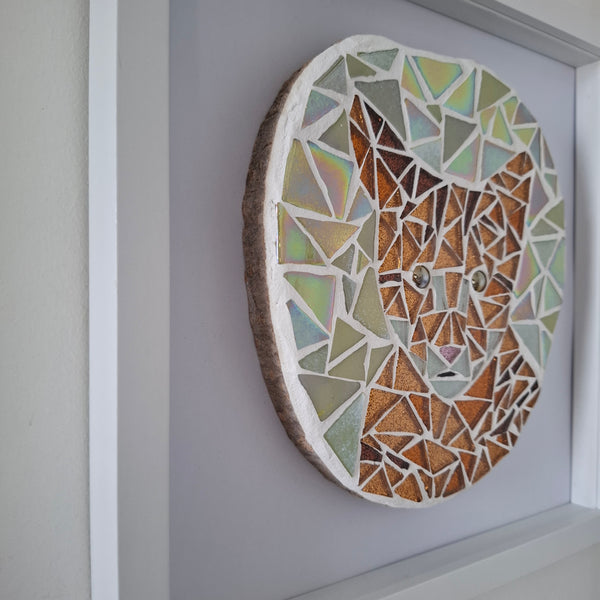 A circular mosaic wall art piece featuring a stylized cat face, mounted on a light gray wall within a white frame. The mosaic is made of geometric pieces in earthy tones of brown, beige, sage green, and iridescent pastel colors.
