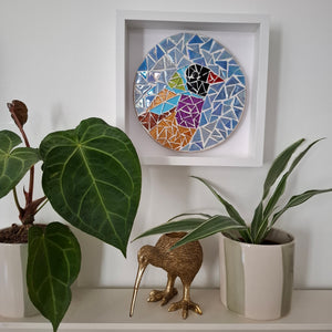 A square white shadow box frame displaying a colorful mosaic bird artwork, styled on a white shelf with potted green plants and a gold kiwi bird figurine.