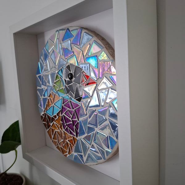 A circular mosaic wall art bird with iridescent glass tiles in blue, purple, pink, orange, and silver, mounted inside a white frame.