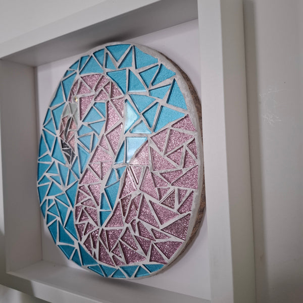 A circular mosaic wall art piece featuring a stylized flamingo, constructed with light blue and pink glitter tiles and white grout, mounted inside a white recessed shelf.