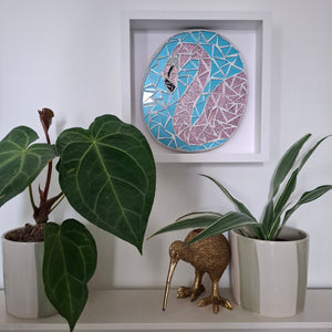 A framed mosaic wall art piece featuring a stylized pink flamingo on a light blue background, displayed on a white shelf above potted plants and a small kiwi figurine.