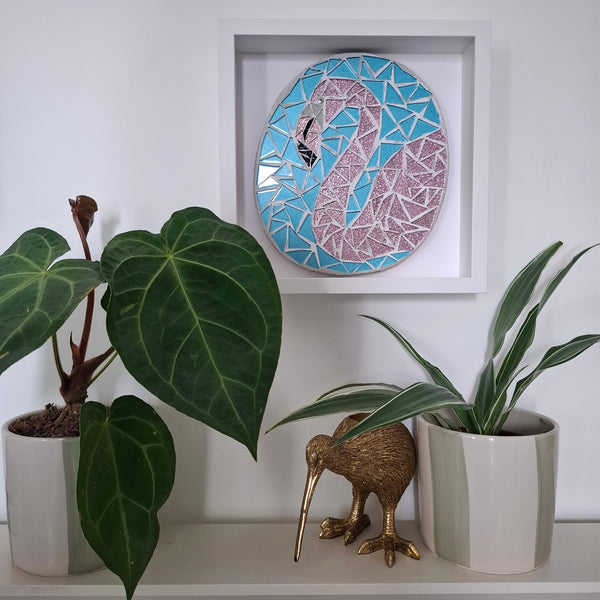 A framed mosaic wall art piece featuring a stylized pink flamingo on a light blue background, displayed on a white shelf above potted plants and a small kiwi figurine.